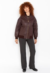 Veste bombardier en cuir marron avec une fermeture éclaire à l'avant, des poignets et un ourlet côtelés. Elle dispose d'une poche latérale avec une fermeture éclair et d'une texture lisse.