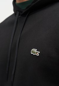 Μαύρο φούτερ με κορδόνια και ένα μικρό πράσινο λογότυπο κροκοδείλου κεντημένο στην αριστερή πλευρά του στήθους.