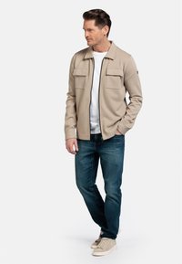 Beige ritsjack met twee borstzakken, gecombineerd met donkerblauwe jeans en lichtbeige sneakers, op een model tegen een witte achtergrond.