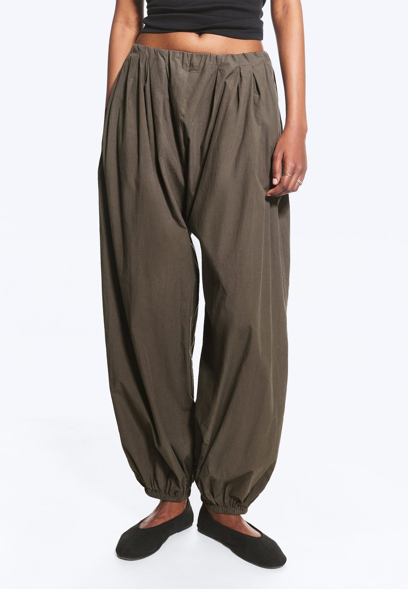 Personne portant un pantalon ample olive foncé avec des poignets élastiques, des ballerines noires et un top court noir, debout devant un fond uni.