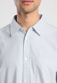 Camicia a righe blu chiaro in cotone. Presenta un colletto button-down, taschino frontale e bottoni bianchi. Texture liscia con linee verticali.
