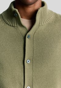 Cardigan en tricot vert olive avec un col côtelé, fermeture par boutons et texture douce. Des boutons bleus remarquables ajoutent un contraste.