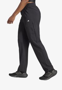 Schwarze Sporthosen mit lockerer Passform und elastischem Bund, ausgestattet mit Seitentaschen und einem Adidas-Logo an der Hüfte. Kombiniert mit schwarzen Laufschuhen.