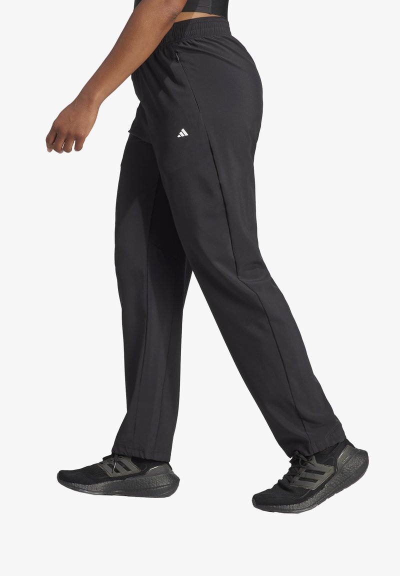 Pantalones deportivos negros de corte holgado y cinturilla elástica, con bolsillos laterales y un logo de Adidas en la cadera. Combinándolo con zapatos de correr negros.