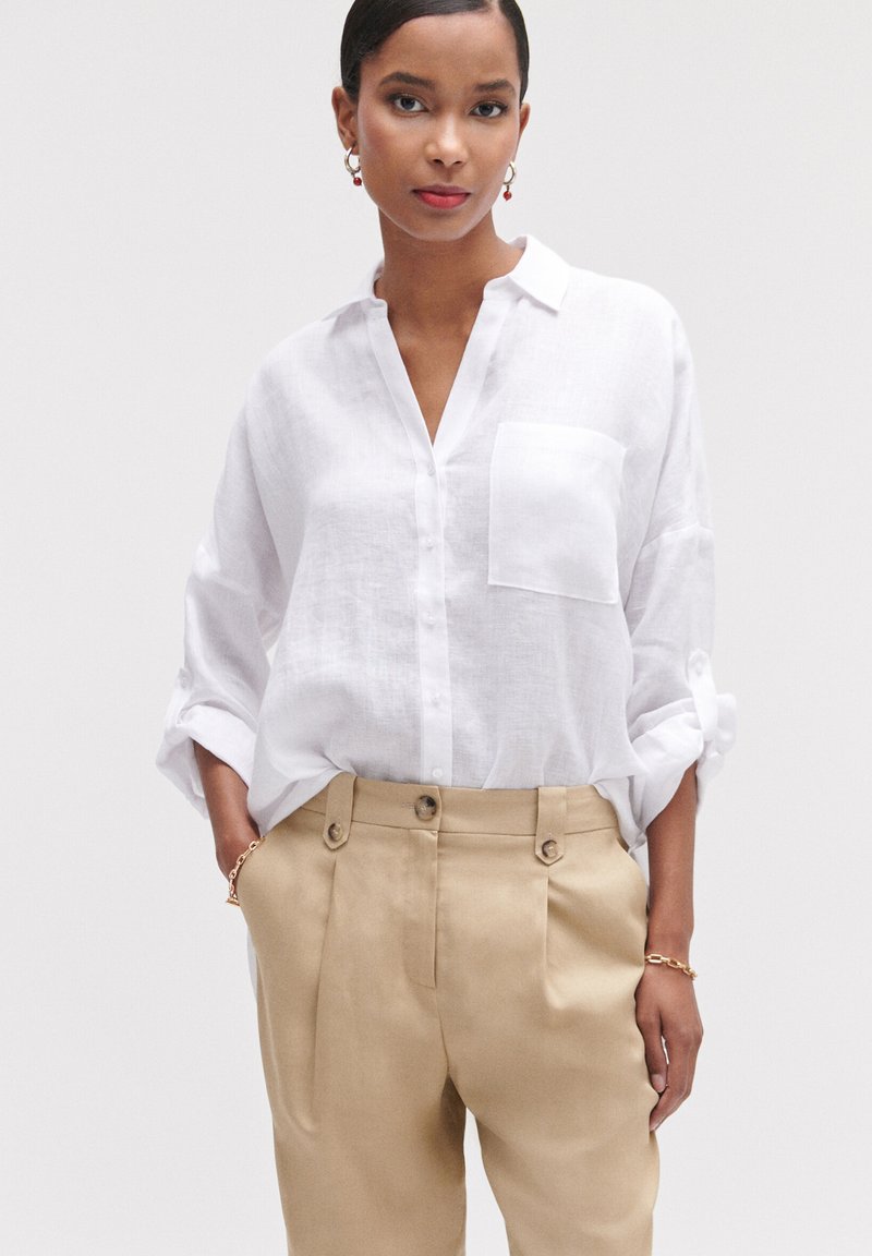 Camicia bianca in lino con taschino sul petto, maniche arrotolate, abbinata a pantaloni beige a vita alta con doppi bottoni.