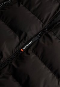 Czarna kurtka puchowa o pikowanej strukturze i widocznym zamku. Posiada etykietę "SUPERDRY" w białym kolorze z pomarańczowym prostokątem.
