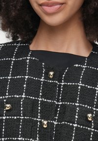 Sort tekstureret jakke med et hvidt rutemønster, der har guldhardware knapper og en rund halsudskæring. Nærbillede med detaljer.