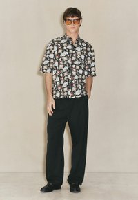 Schwarzes Kurzarmhemd mit buntem Cartoon-Muster, kombiniert mit lockeren schwarzen Hosen und schwarzen Schuhen. Braune Sonnenbrillen runden den Look ab.