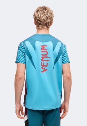 Personne aux cheveux blonds courts portant un maillot de sport bleu Venum avec un logo rouge et des manches à motifs, vue de dos sur un fond uni.