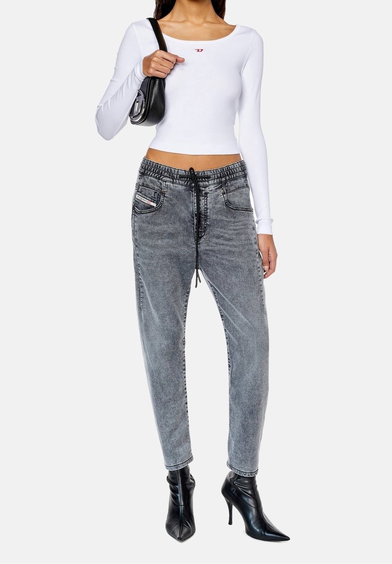 Top corto de manga larga blanca con un pequeño logo rojo, combinado con jeans gris de estilo jogger con cordón y botas de tacón negro de punta.