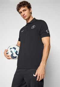 Sort polo med krave, logo på brystet og Nike-design elementer. Modellen holder en hvid og blå fodbold.