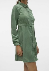 Žena v zelených manšestrových šatech s dlouhými rukávy, zapínáním na knoflíky, límečkem a zavázaným páskem, zobrazená od poloviny stehna po krk.