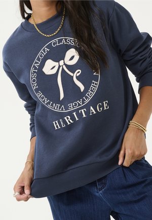 Person iført en mørkeblå sweatshirt med en hvid sløjfe-grafik og cirkulær tekst, der lyder "Classic Nostalgia Heritage Vintage."