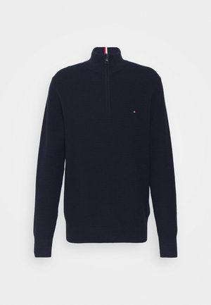 Pull tricot bleu marine à fermeture éclair partielle avec manches longues et col montant, avec un petit logo rouge, blanc et bleu sur la poitrine.