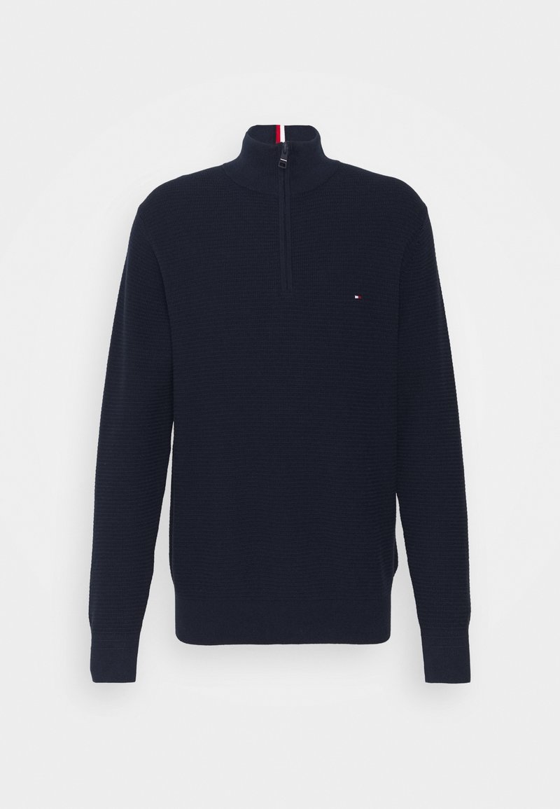 Pull tricot bleu marine à fermeture éclair partielle avec manches longues et col montant, avec un petit logo rouge, blanc et bleu sur la poitrine.