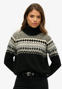 Schwarzer gestrickter Pullover mit hohem Kragen, versehen mit einem gemusterten, hellgrauen und cremefarbenen Schulterbereich. Akzentuiert mit einem schwarzen Gürtel und dunklen Jeans.