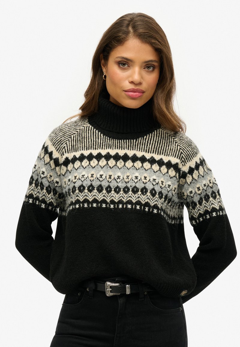 Schwarzer gestrickter Pullover mit hohem Kragen, versehen mit einem gemusterten, hellgrauen und cremefarbenen Schulterbereich. Akzentuiert mit einem schwarzen Gürtel und dunklen Jeans.