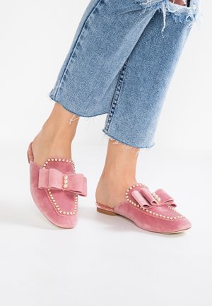 Pieds portant des chaussures en velours rose à enfiler, ornées de perles et de grands nœuds, associées à un jean court bleu clair à ourlet effiloché, debout sur un sol blanc.