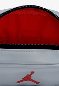 Borsetta grigia con fodera interna rossa, cerniera nera e logo Jumpman rosso. Texture esterna liscia, forma rettangolare e senza ulteriori motivi.