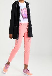 Musta huppari, valkoinen graafinen t-paita, vaaleanpunaiset farkut ja purppurat lenkkarit oransseilla yksityiskohdilla; rento asu monilla väreillä ja tekstuureilla.