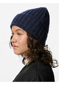 Mörkblå stickad mössa med ribbstruktur, avsmalnande form och mjukt tyg. Buren med en svart quiltad jacka, vilket visar en sidoprofil.