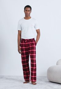 Άσπρο κοντομάνικο πουκάμισο με το λογότυπο "HUGO", κόκκινο καρό παντελόνι πυτζάμας με μαύρες και λευκές λεπτομέρειες, χαλαρή εφαρμογή, φορεμένο ξυπόλυτο.