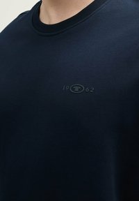 Navy Crew-Neck Sweatshirt mit weichem Material, geripptem Kragen und einem dezenten geprägten Logo mit dem Jahr "1962" auf der Brust.