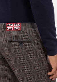 Wool blend trousers featuring a brown and blue check pattern, a single back pocket with a navy button, and a label with a Union Jack design.