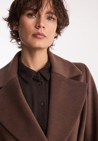 Brązowy strukturalny płaszcz z szerokim kołnierzem, teksturowana tkanina oraz dopasowana ciemna koszula na guziki pod spodem. Zbliżenie bez szczegółów tła.