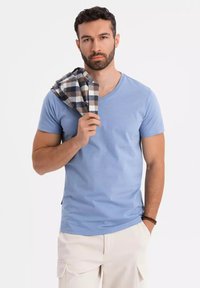 Světle modré bavlněné tričko s výstřihem do V, krátkými rukávy; modelka drží kostkovaný šedo-hnědý šál na bílém pozadí.