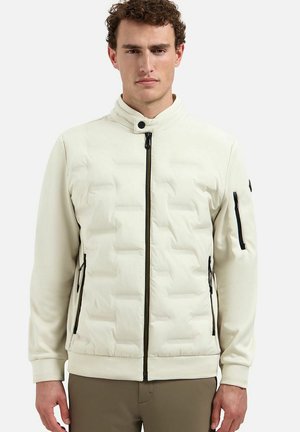 Man met krullend haar die een lichtbeige doorgestikte jas met zwarte rits draagt en staat tegen een effen lichte achtergrond.