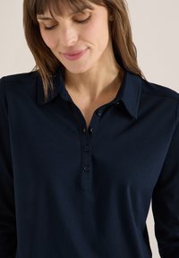 Femme aux cheveux bruns souriant doucement, portant une chemise bleu marine à manches longues avec col et quatre boutons sur la patte de boutonnage.