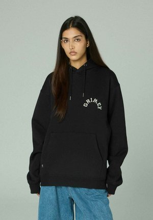 Jonge vrouw met lang donker haar draagt een zwarte hoodie met de tekst "GRIMEY" en losse blauwe spijkerbroek, staand tegen een eenvoudige lichte achtergrond.