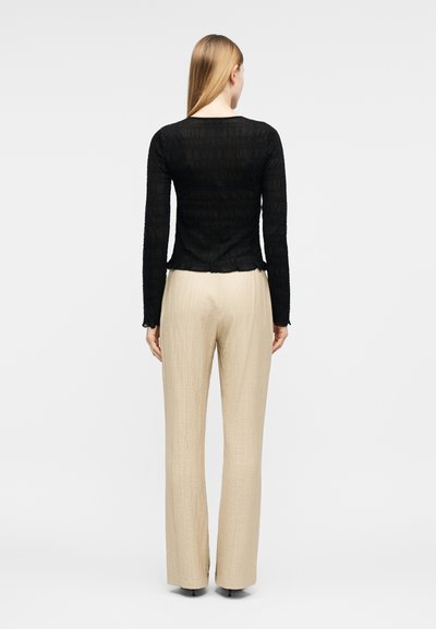 Γυναίκα με μακριά ξανθά μαλλιά που φοράει μαύρο υφασμάτινο τοπ με μακριά μανίκια και μπεζ παντελόνι φαρδιάς γραμμής, όρθια με την πλάτη προς τα εμάς σε λευκό φόντο.