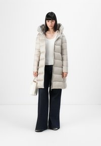 Lichtbeige gewatteerde jas met capuchon, voorzien van een geborduurd ontwerp, een rits aan de voorkant en een zijzak met rits. Gestyled met vrouwelijke donkere spijkerbroek met flare.