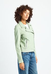 Miętowo-zielona skórzana kurtka motocyklowa z zamkami błyskawicznymi w kieszeniach, kołnierzem na zatrzaski i kontrastowym przeszyciem. Noszona na białej koszuli i niebieskich dżinsach.