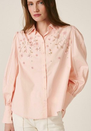 Mujer con una blusa de botones de color rosa claro, con bordados florales y detalles de cuentas en el pecho y mangas largas abullonadas.