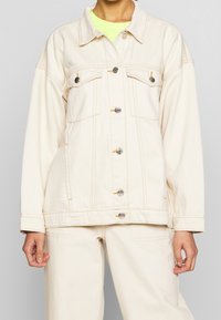 Beige denimjacka med knäppning framtill, två bröstfickor och avslappnad passform. Har silverfärgade knappar och synliga sömsdetaljer.