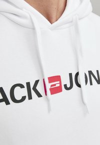 Hvit hettegenser med svart "JACK&JONES"-tekst og en rosa bokslogo. Har snøring og mykt, teksturert stoff.