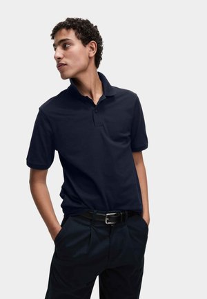 Jonge man met krullend haar, gekleed in een donkerblauw poloshirt en bijpassende broek, met zijn handen in zijn zakken, kijkt naar links.