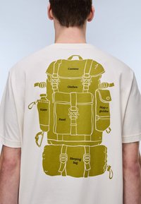 Ανοιχτόχρωμο μπλουζάκι με γράφημα ενός σακιδίου πλάτης σε ελιά πράσινο, με ετικέτες αντικειμένων: φωτογραφική μηχανή, ρούχα, νερό, φαγητό, χάρτες, υπνόσακος.