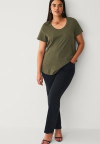 Oliven grøn t-shirt med rund hals, kombineret med mørke tætsiddende jeans. Modellen står med hænderne i lommerne iført sorte sandaletter med stropper.