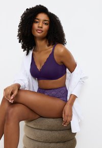 Purppura pitsivaatteet, joissa on V-kaula-aukko ja huipulla reunat, sekä korkeavyötäröiset alushousut, joita käytetään avonaisen valkoisen paidan alla.