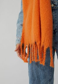 Personne portant une veste en jean bleu clair et un jean avec une écharpe en laine orange vif à franges pendante à côté de sa main.