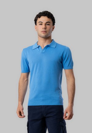 Giovane uomo in piedi che indossa una polo azzurro chiaro a maniche corte e pantaloni cargo scuri su uno sfondo grigio uniforme.