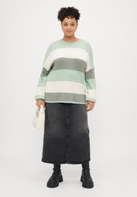 Ombre zelene, bele in sive črtaste prevelike pulover, v kombinaciji z dolgim črnim denim krilom in črnimi robustnimi čevlji, drži majhno kremo torbico.