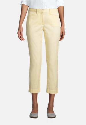 Femme portant un pantalon court jaune clair, une chemise blanche à boutons aux manches retroussées et des ballerines grises, debout devant un fond uni.