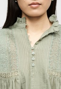 Κοντινό πλάνο ενός ατόμου που φοράει ανοιχτοπράσινο πουκάμισο με δαντελένιες λεπτομέρειες, κουμπιά και λαιμόκοψη με βολάν, εμφανίζοντας την κλείδα και τα χείλη.