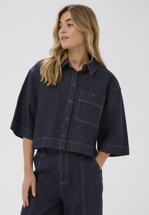 Blonde woman wearing dark blue cropped denim shirt with buttoned chest pocket and matching high-waisted denim pants.