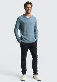 Lächelnder Mann mit braunem Haar, der einen hellblauen V-Ausschnitt-Pullover, dunkle Hosen und weiße Turnschuhe trägt, steht mit den Händen in den Taschen.